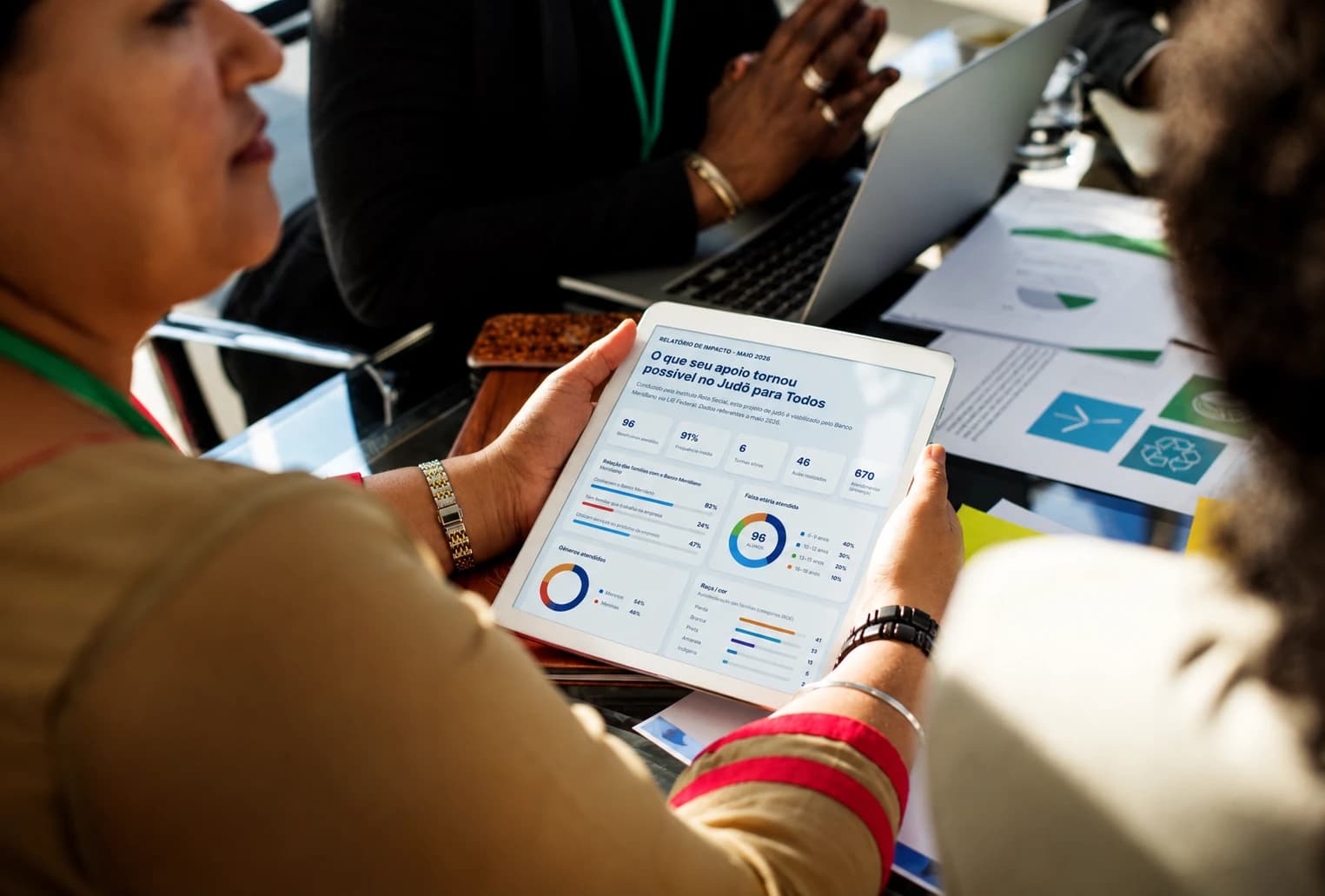 Duas mulheres em uma reunião analisando em um tablet o relatório de impacto de um projeto apoiado, com gráficos e indicadores na tela